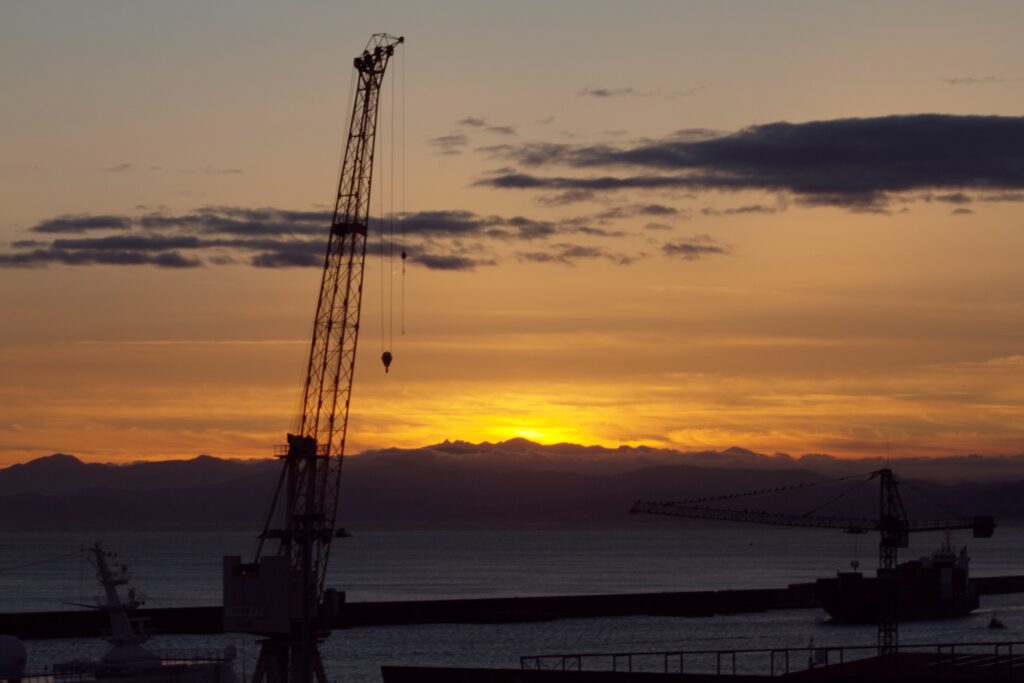 Tramonto sul porto di Genova con la silhouette delle gru portuali e il sole che cala sulle montagne