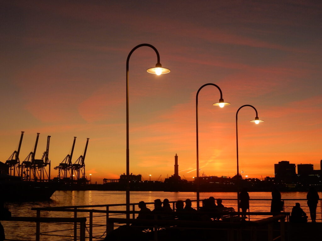 People walking on a Genoese pier at sunset, with lit lamps and harbour cranes silhouetted